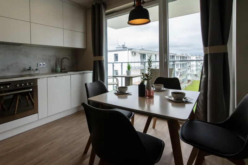 wnętrze apartamentu, widok na aneks kuchenny, stół jadalny oraz drzwi balkonowe z widokiem na nowoczesne osiedle