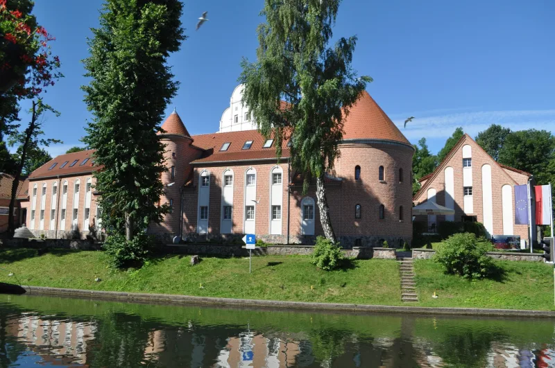 Widok na hotel, przed nim dwa wysokie drzewa i woda w kanale