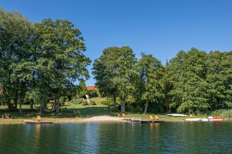 zdjęcie na plażę z pomostami, na brzegu leżakami i kajakami. W głębi dach domostwa pomiędzy drzewami.